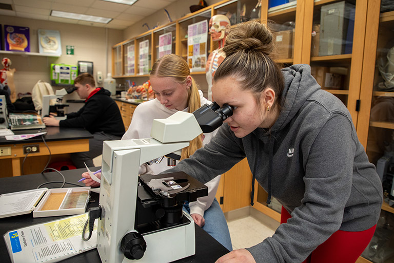 two female students in a biology lab with one looking in a microscope